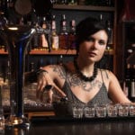 A female bartender standing behind a bar, pouring a clear liquor into six shot glasses lined up in front of her, and shelves of liquor bottles in the background.