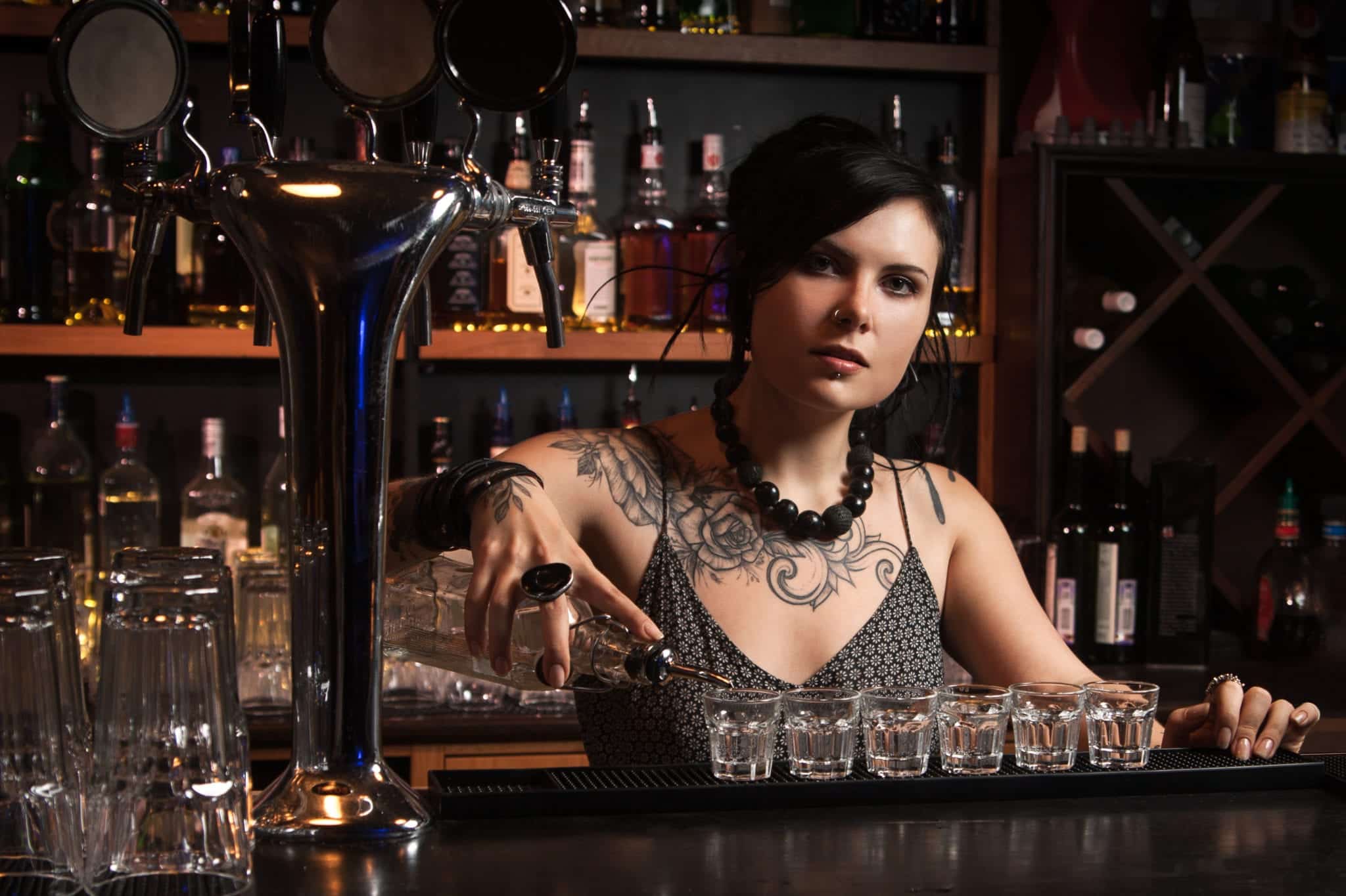 A female bartender standing behind a bar, pouring a clear liquor into six shot glasses lined up in front of her, and shelves of liquor bottles in the background.