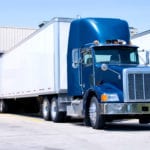 A blue tractor trailer with a white trailer backed up at a warehouse loading dock.