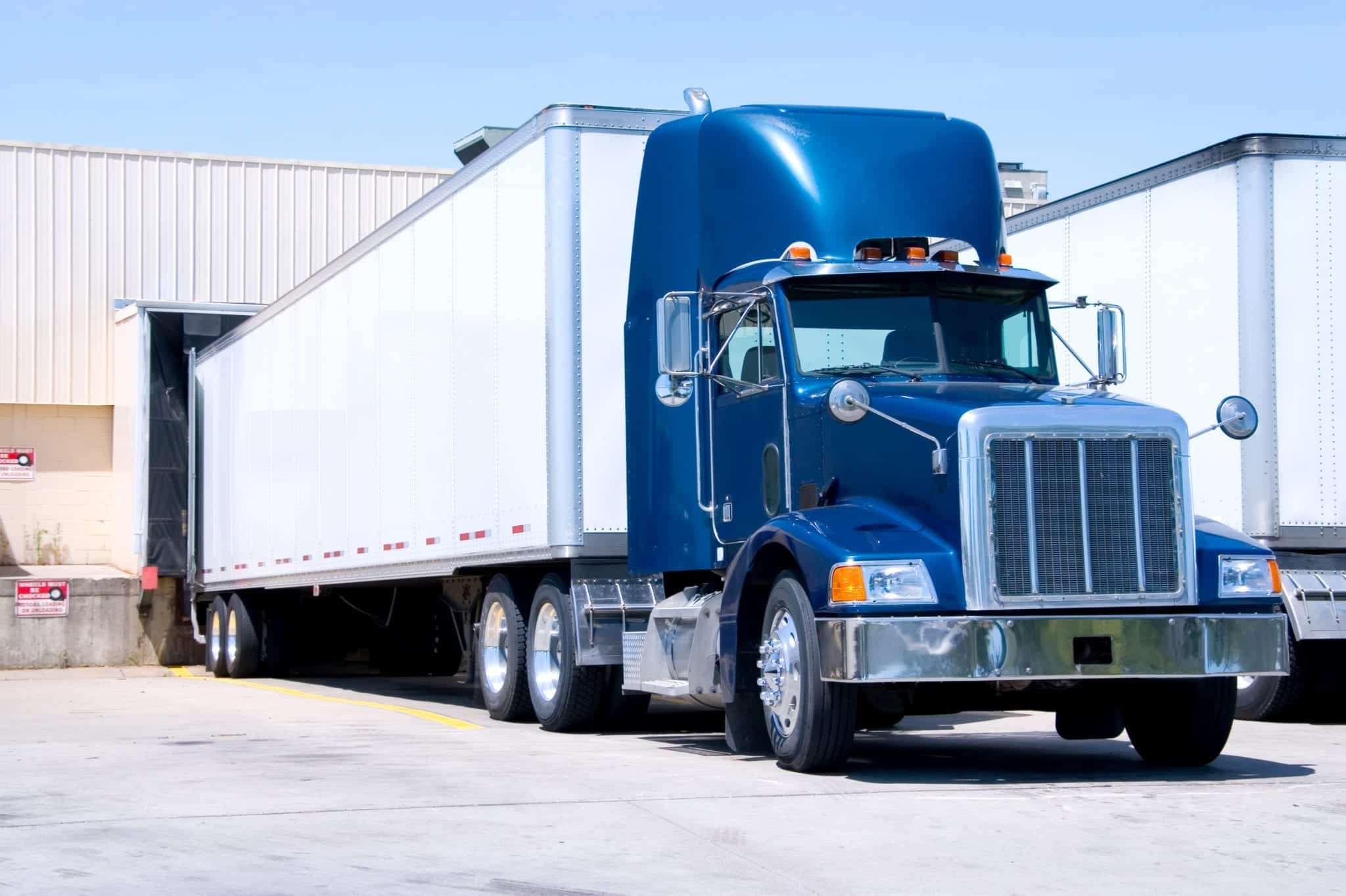 A blue tractor trailer with a white trailer backed up at a warehouse loading dock.