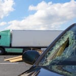 In the foreground is a car with a large hole smashed through the windshield, a pile of wood and a tractor trailer in the background.