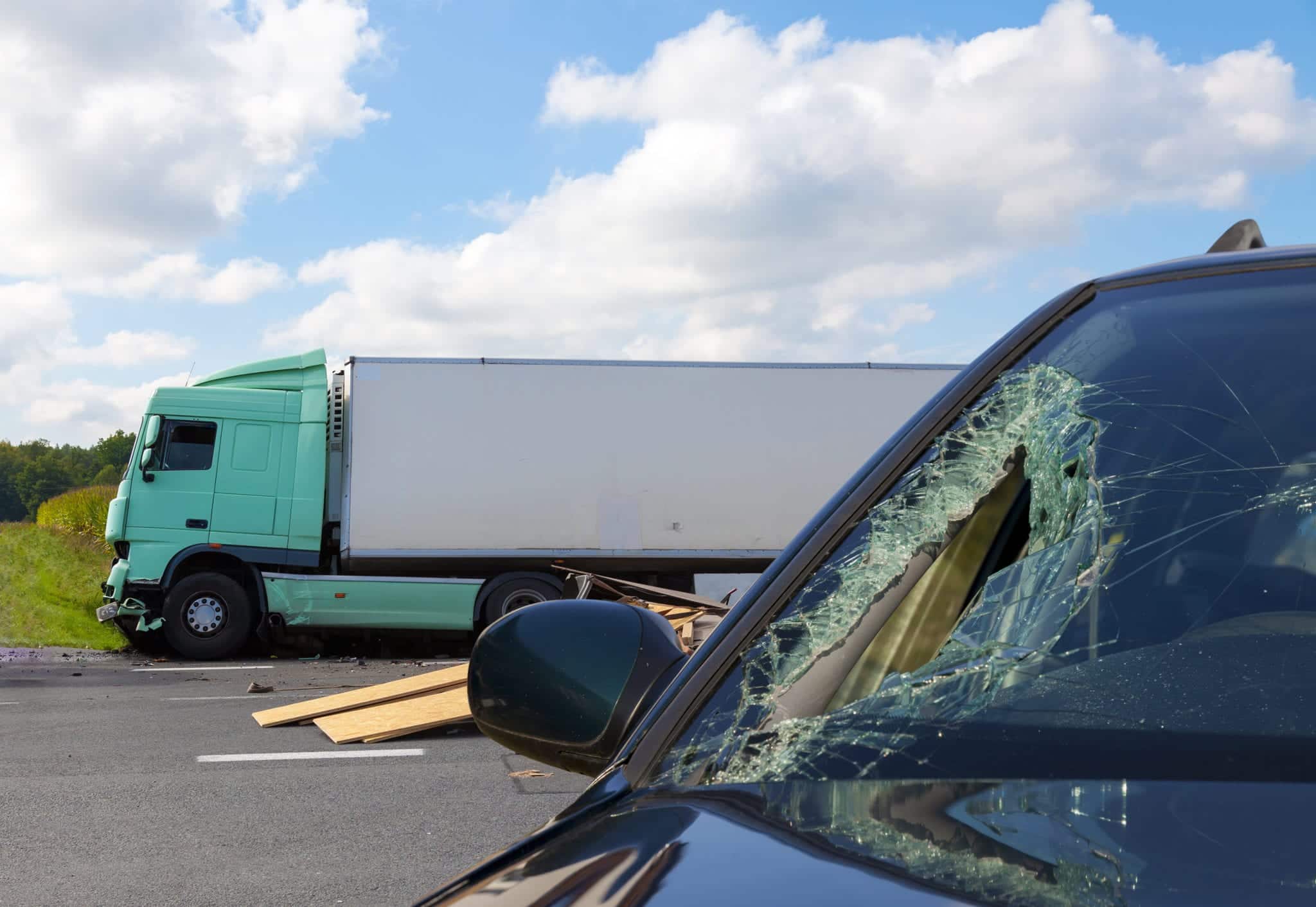 In the foreground is a car with a large hole smashed through the windshield, a pile of wood and a tractor trailer in the background.