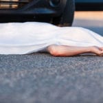 A white sheet covering everything except the left arm of a dead body, laying on a road, in front with a black car.