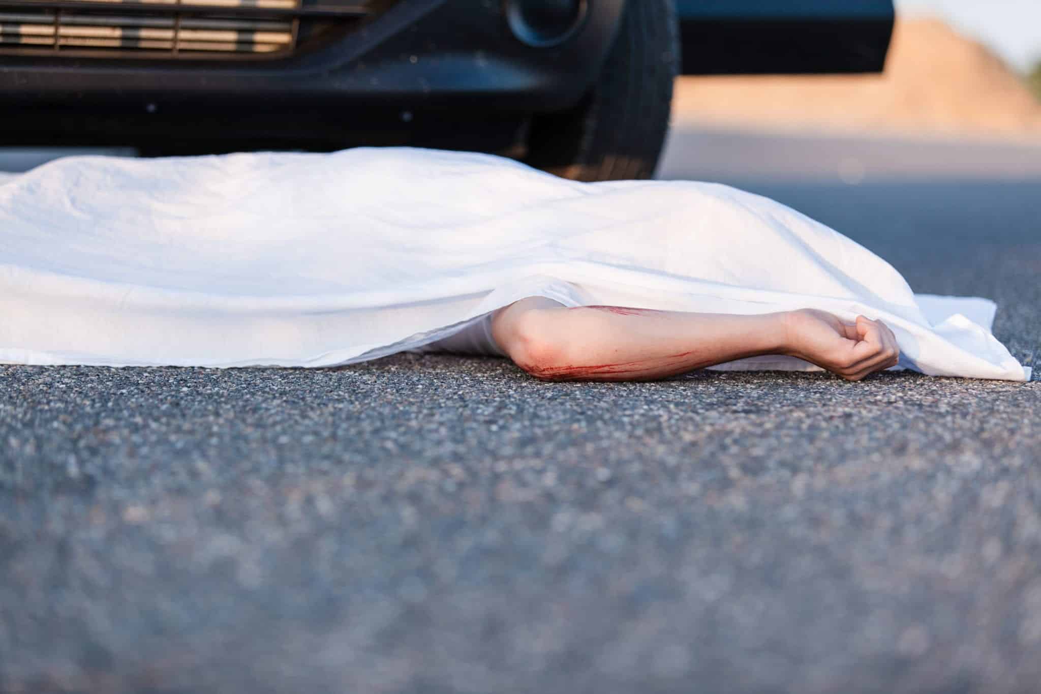 A white sheet covering everything except the left arm of a dead body, laying on a road, in front with a black car.