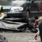 The scene of an auto accident with an injured man kneeling down in the foreground and a paramedic zipping up a black body bag in the background