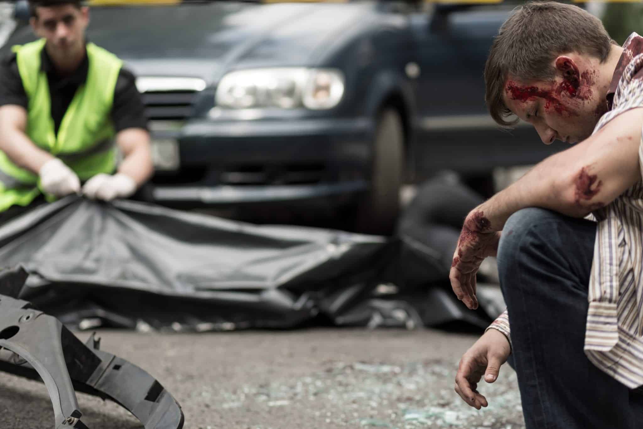 The scene of an auto accident with an injured man kneeling down in the foreground and a paramedic zipping up a black body bag in the background