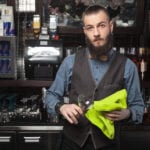 A male bartender, standing in front of a counter and shelves with bar glassware, using a neon green towel to dry a wine glass.