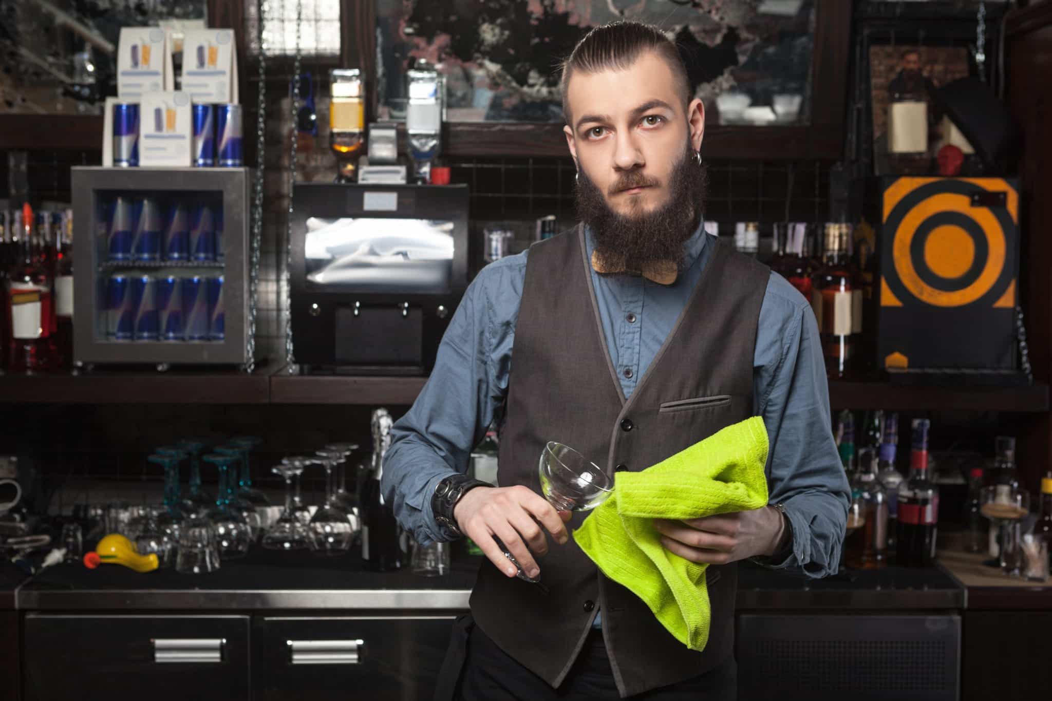 A male bartender, standing in front of a counter and shelves with bar glassware, using a neon green towel to dry a wine glass.