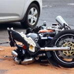 A wrecked motorcycle laid in the road with a silver car in the background.