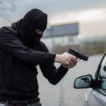 An armed, masked man, wearing all black, pointing a gun at a car window.