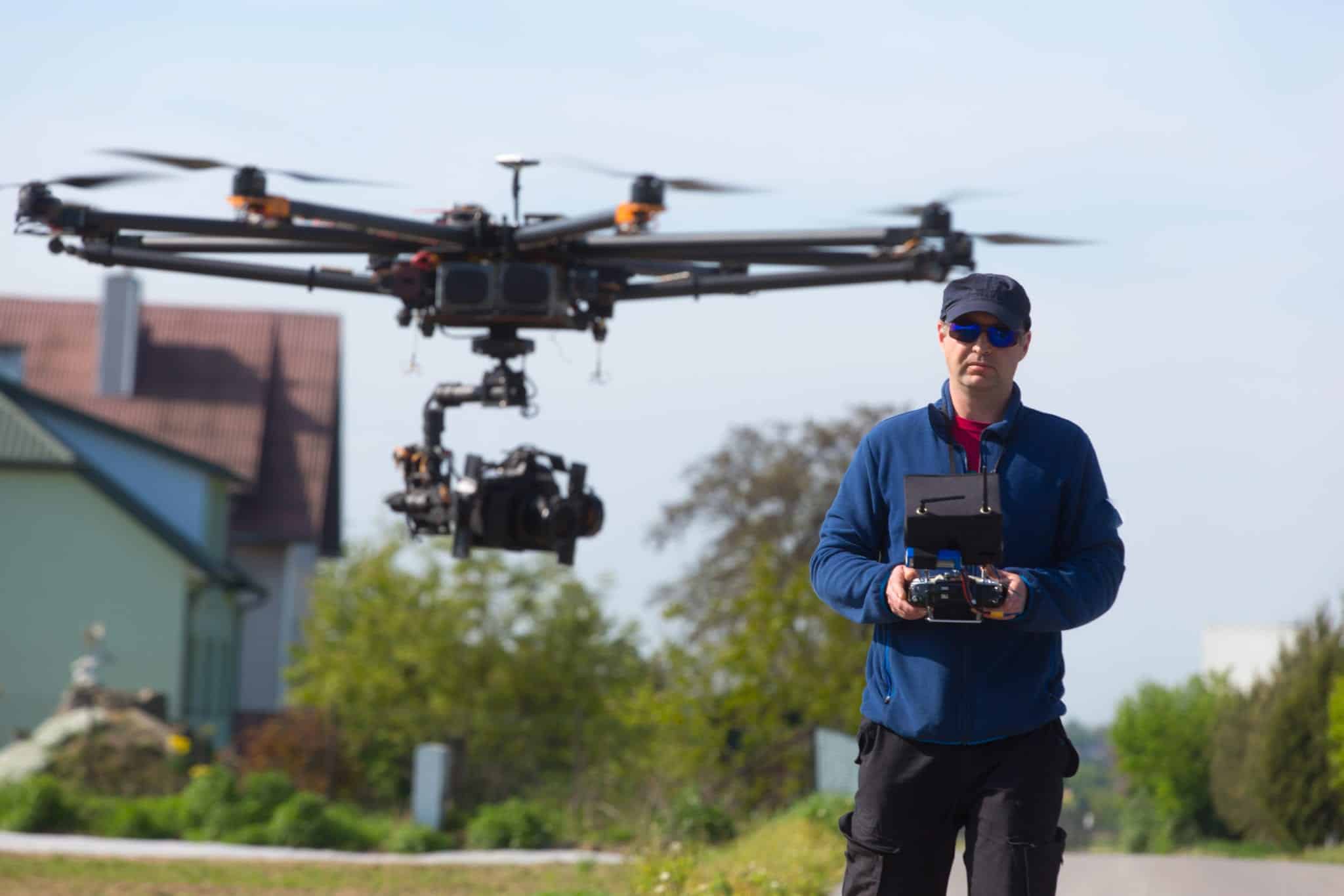 A man wearing a navy blue jacket, hat, sunglasses and black pants flying a large drown scene in the foreground with a blurred image of a house and trees in the background.