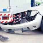 The front of a silver truck with it's bumper completely off next to the back end of a tractor trailer that appears to have hit the truck.