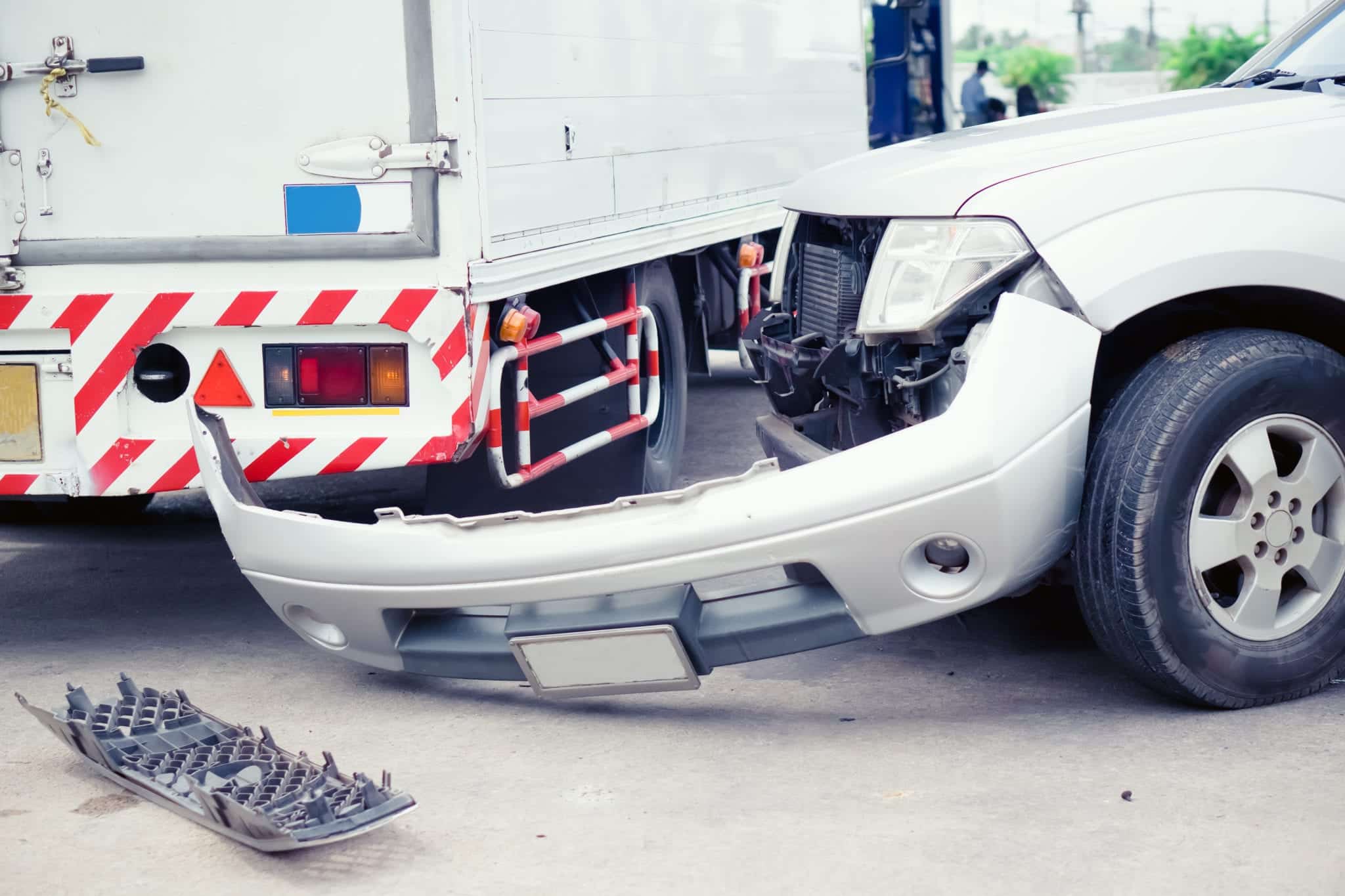 The front of a silver truck with it's bumper completely off next to the back end of a tractor trailer that appears to have hit the truck.