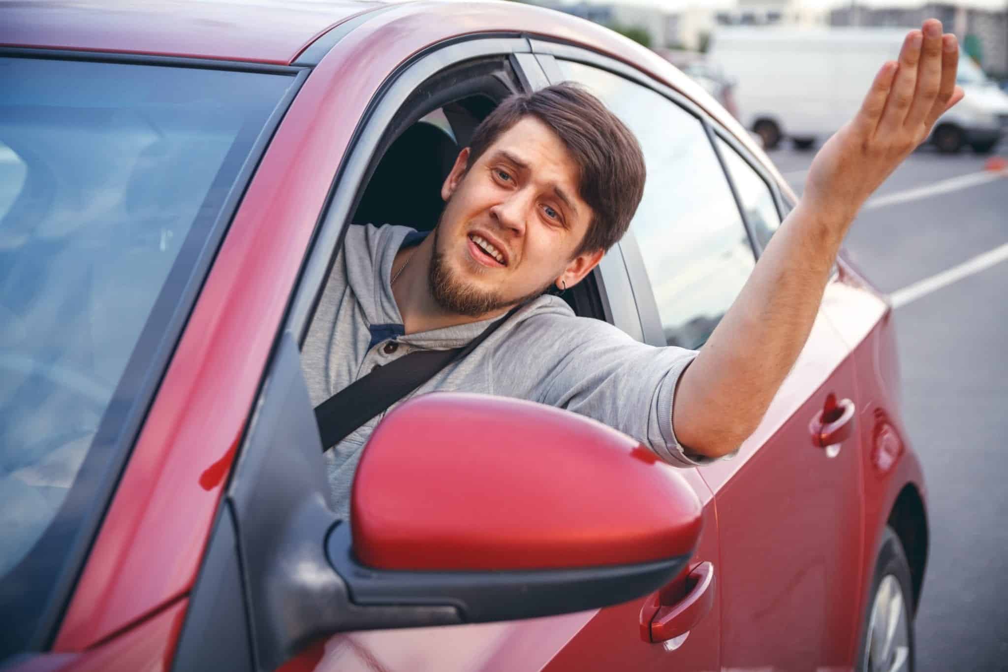 A man looking irritate while sticking his arm and head out of the drivers side window of a small red car.