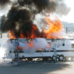 An RV parked near other RVs in a parking lot, with an electrical hook-up, on fire with bright orange flames and dark smoke rising out of it.