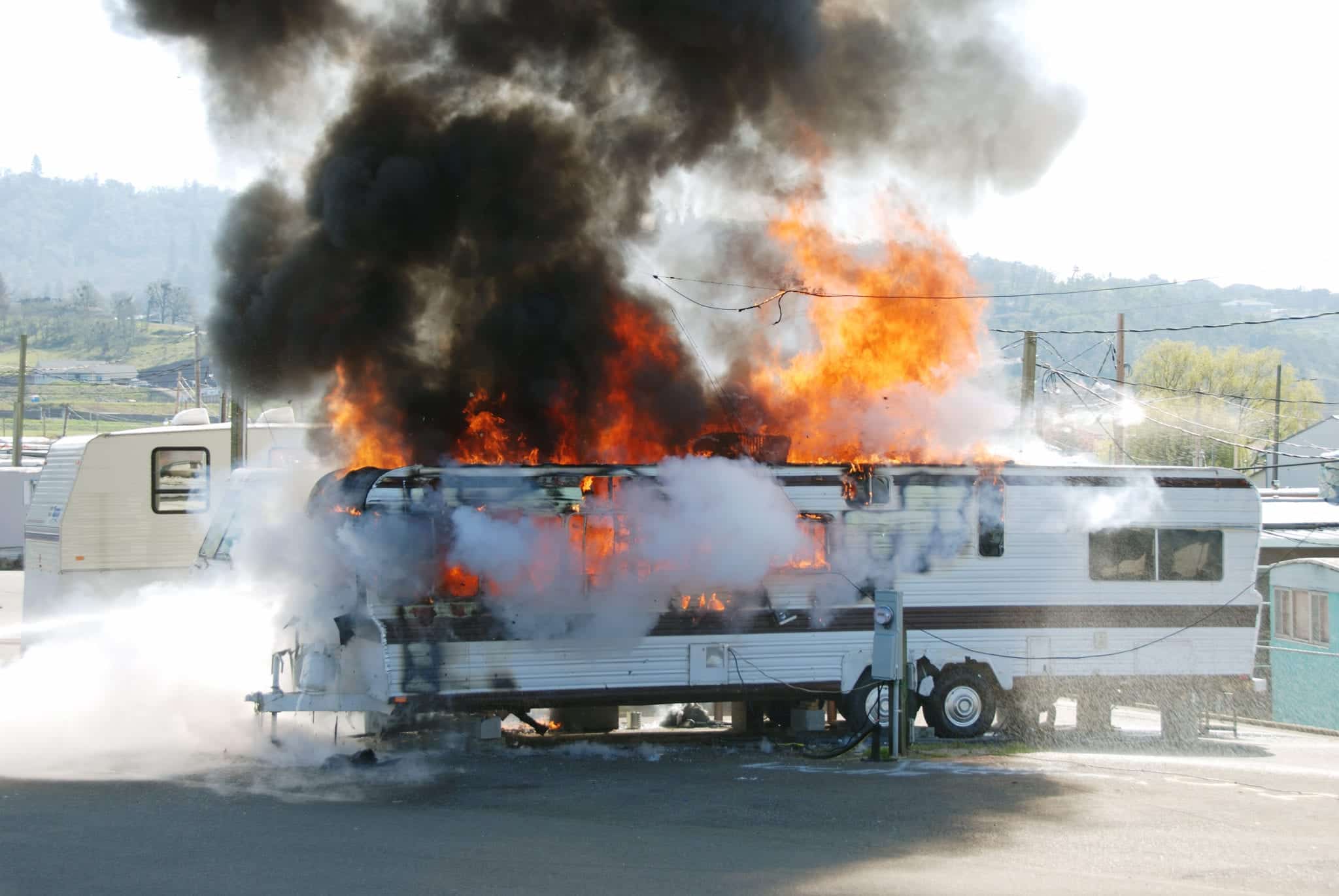 An RV parked near other RVs in a parking lot, with an electrical hook-up, on fire with bright orange flames and dark smoke rising out of it.