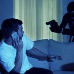 A man wearing a white t-shirt, sitting on a gray couch in a dimly lit room with closed currents in the background being held up at gun point by two individuals disguised in all black.