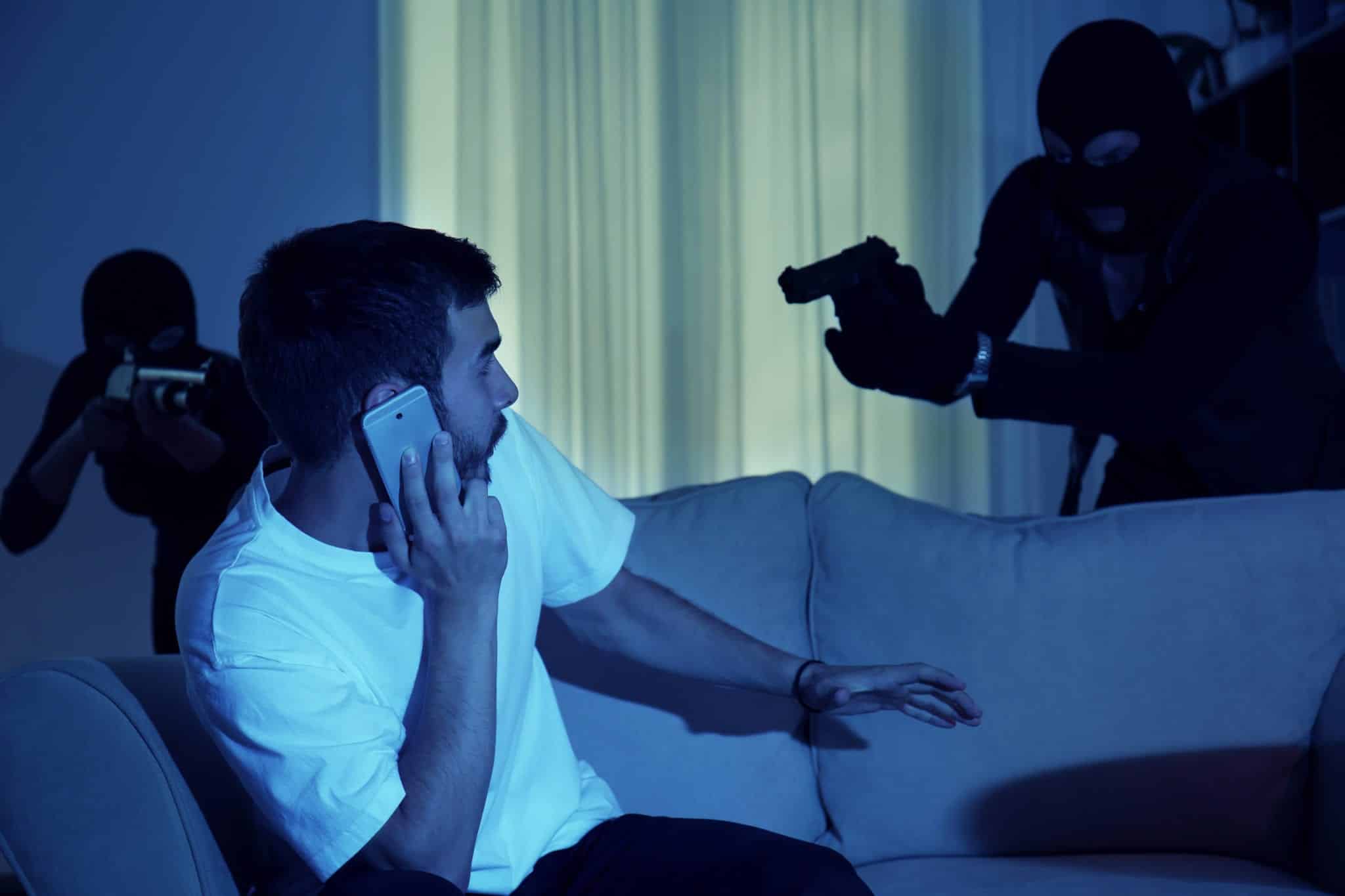 A man wearing a white t-shirt, sitting on a gray couch in a dimly lit room with closed currents in the background being held up at gun point by two individuals disguised in all black.