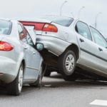 The scene of a wreck on a highway and traffic shown in the blurry background with a silver car having driven into another silver car and pushing the back end of that car up onto the hood of their car.