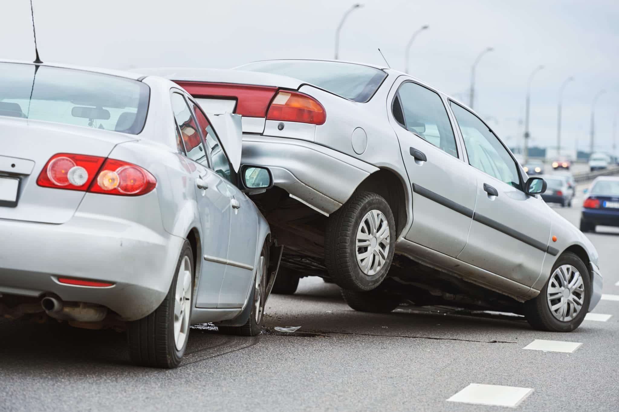 The scene of a wreck on a highway and traffic shown in the blurry background with a silver car having driven into another silver car and pushing the back end of that car up onto the hood of their car.