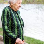 An elderly woman with short white curly hair, a green and black patterned sweater, and black pants, using a brown cane stands near a lake, watching the water.