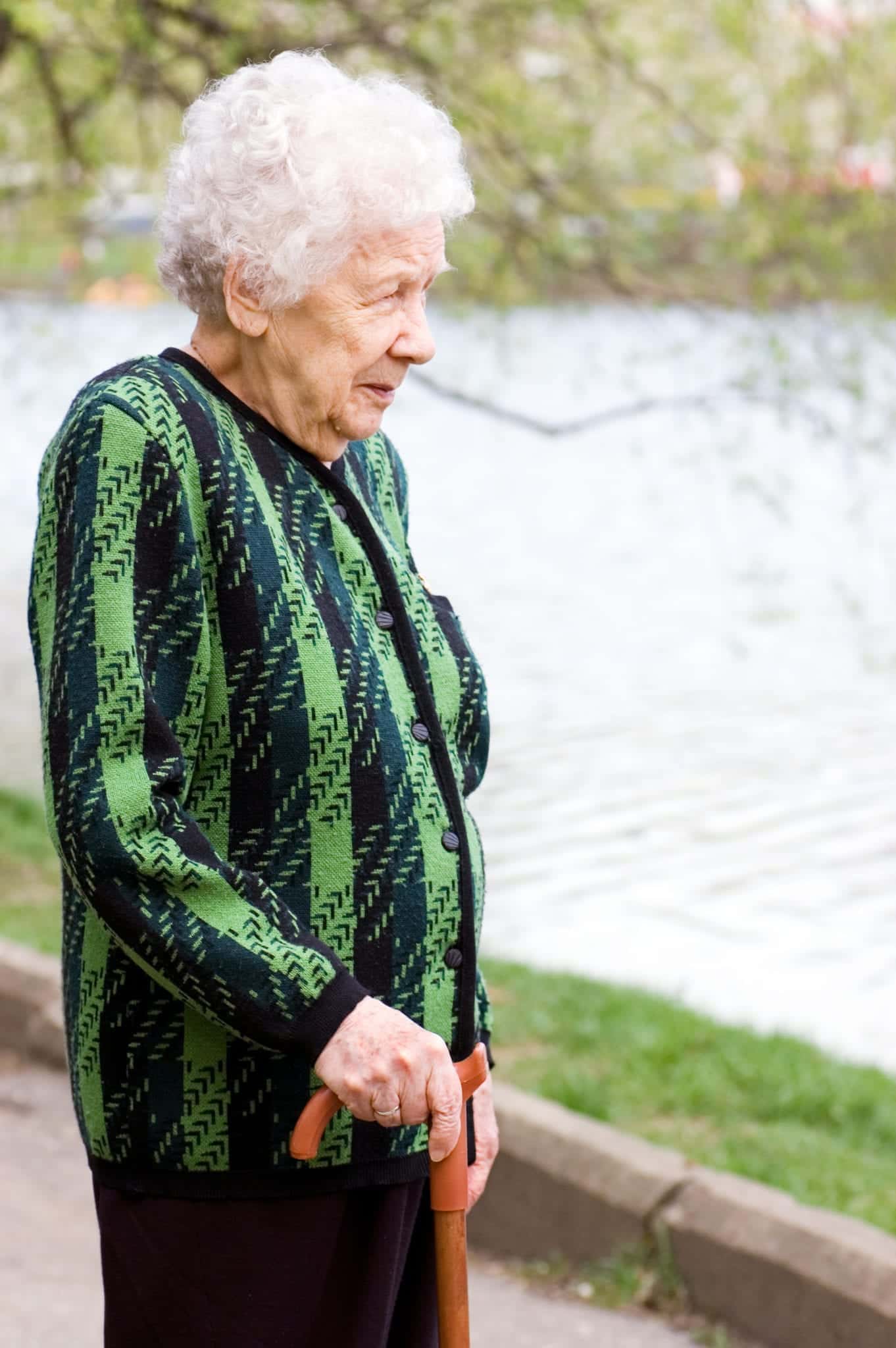 An elderly woman with short white curly hair, a green and black patterned sweater, and black pants, using a brown cane stands near a lake, watching the water.