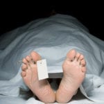 A body laying in the morge with the bottom of that individual's feet seen sticking out from under a white sheet with a white paper tag tied around the right big toe.