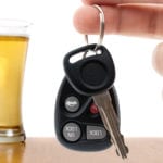 A man's hand holding car keys in the foreground with a full glass of beer on a brown table against a white background.