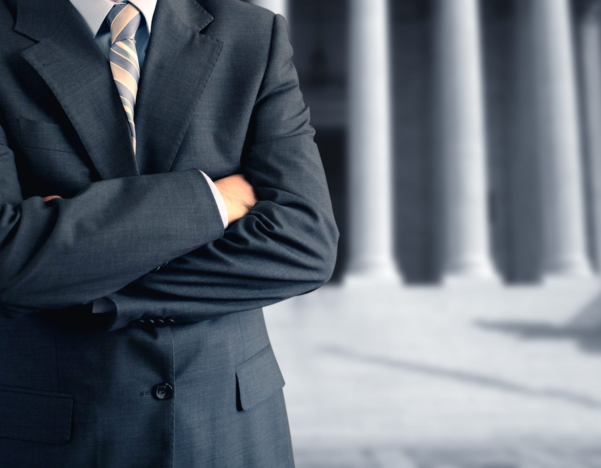 A businessman wearing a black suit with his arms crossed, standing in front of a building with columns.