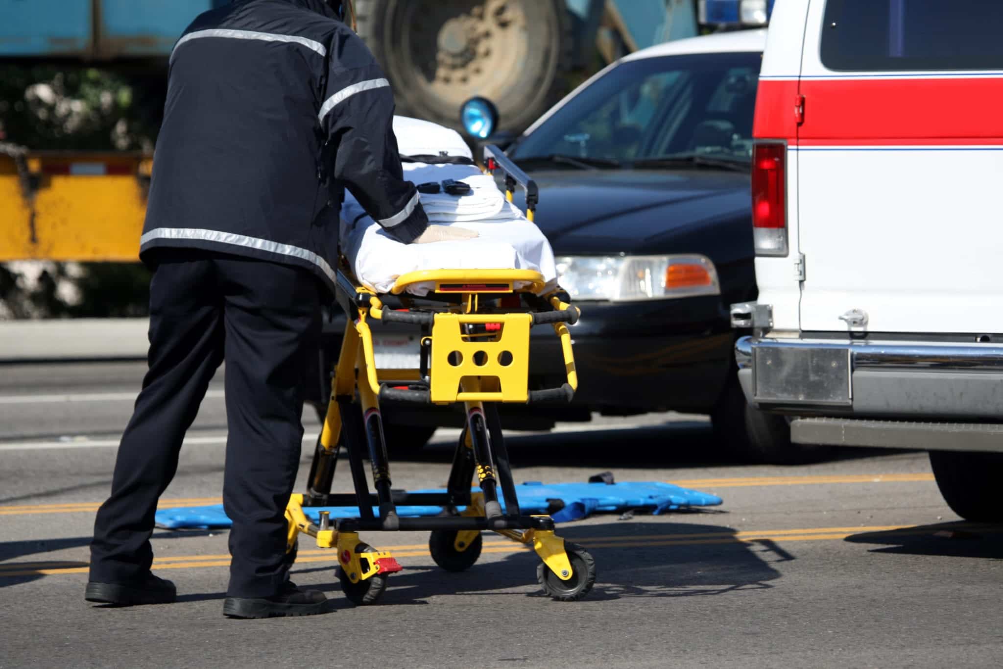 As empty stretcher being rolled by a paramedic on a street near a cop car and an ambulance.