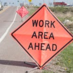 A road with trucks driving towards the viewer with orange signs on the side of the road stating there is a WORK AREA AHEAD and another sign in the background saying BE PREPARED TO STOP indicating a construction zone ahead.