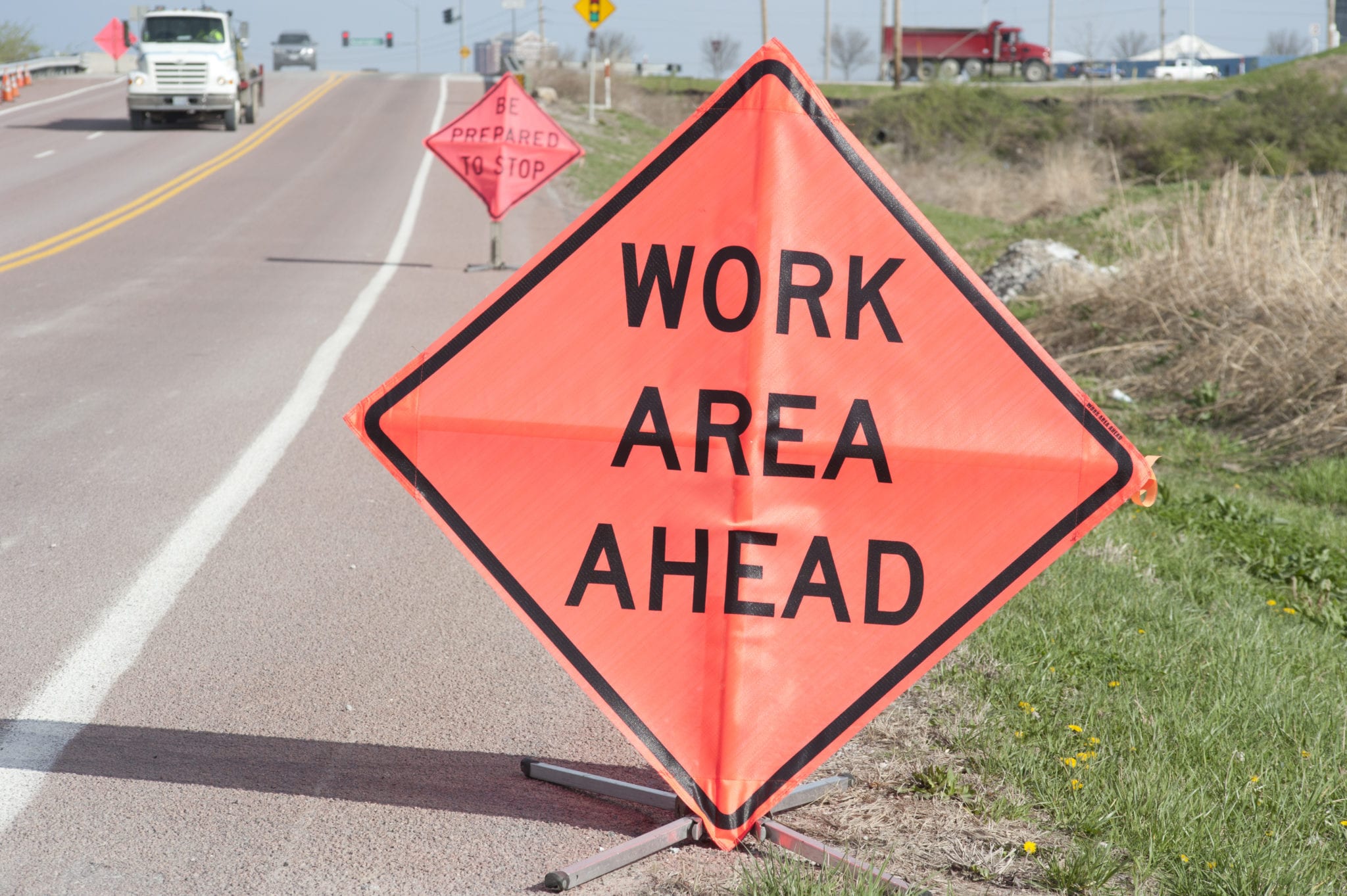 A road with trucks driving towards the viewer with orange signs on the side of the road stating there is a WORK AREA AHEAD and another sign in the background saying BE PREPARED TO STOP indicating a construction zone ahead.