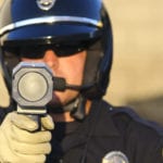 A police officer wearing a black helmet and sunglasses, holding a radar speed detection gun towards the camera.