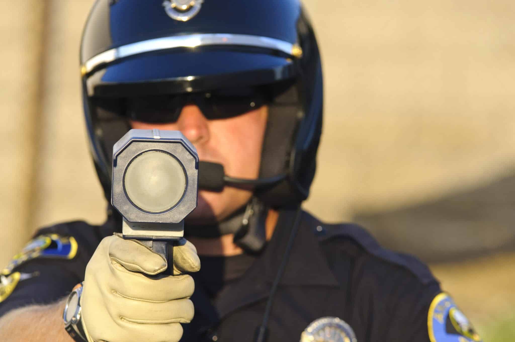 A police officer wearing a black helmet and sunglasses, holding a radar speed detection gun towards the camera.