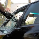 A person wearing a long sleeved dark brown shirt, punching through the window of a black car appearing to have the intent to steal the car.