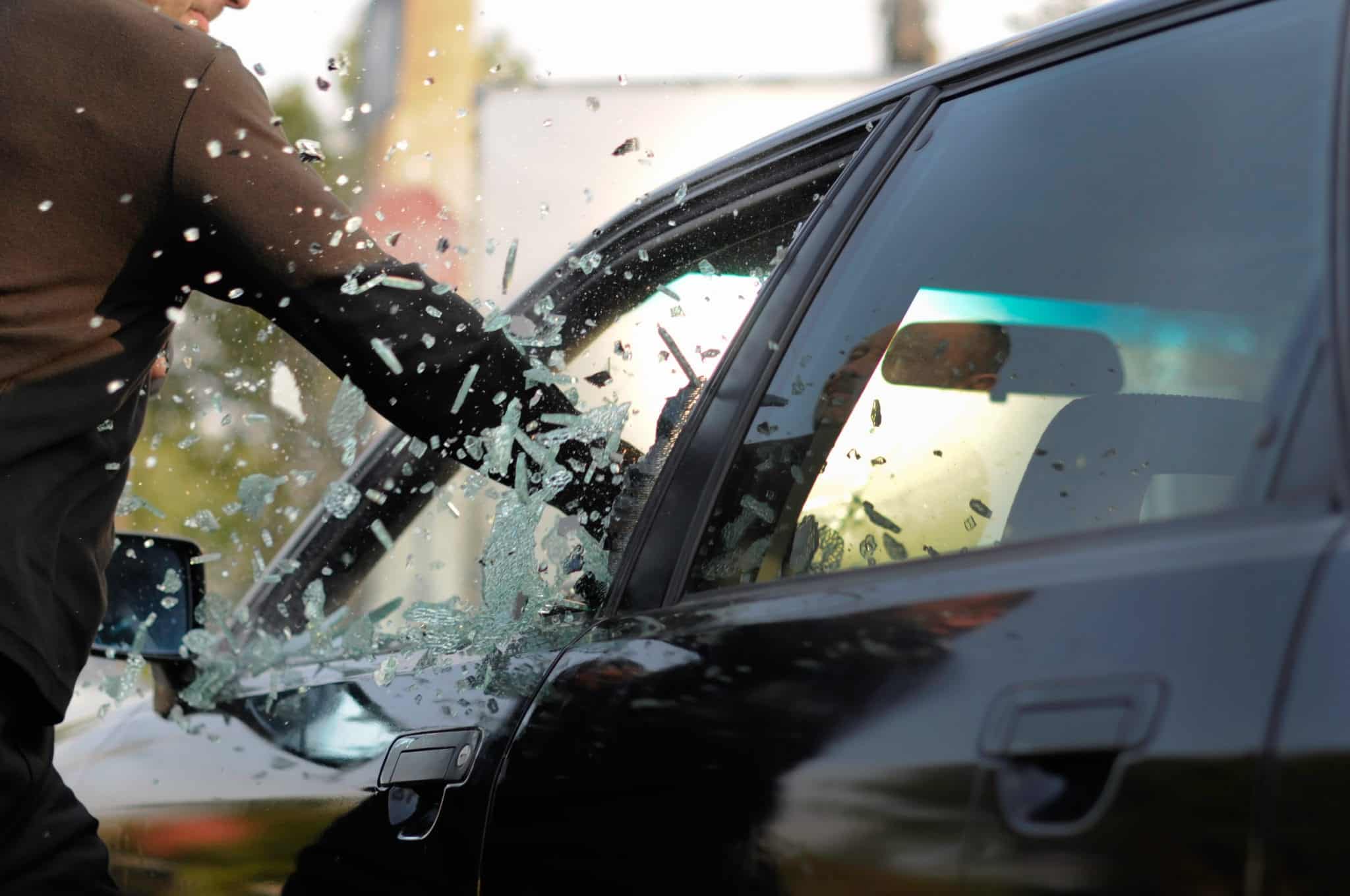 A person wearing a long sleeved dark brown shirt, punching through the window of a black car appearing to have the intent to steal the car.