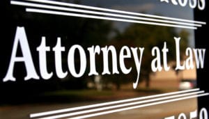 A black sign with the words Attorney at Law in white letters and a blurry scene of an outdoor setting reflecting off of the black sign.