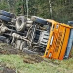 The bottom of a yellow school bus that had rolled off the road onto it's side in the grass, with woods in the background.