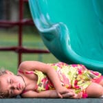 A little girl lying unconscious ground at the bottom of a green slide.