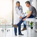 A male patient sitting on an exam table while a older male doctor examines his knee.