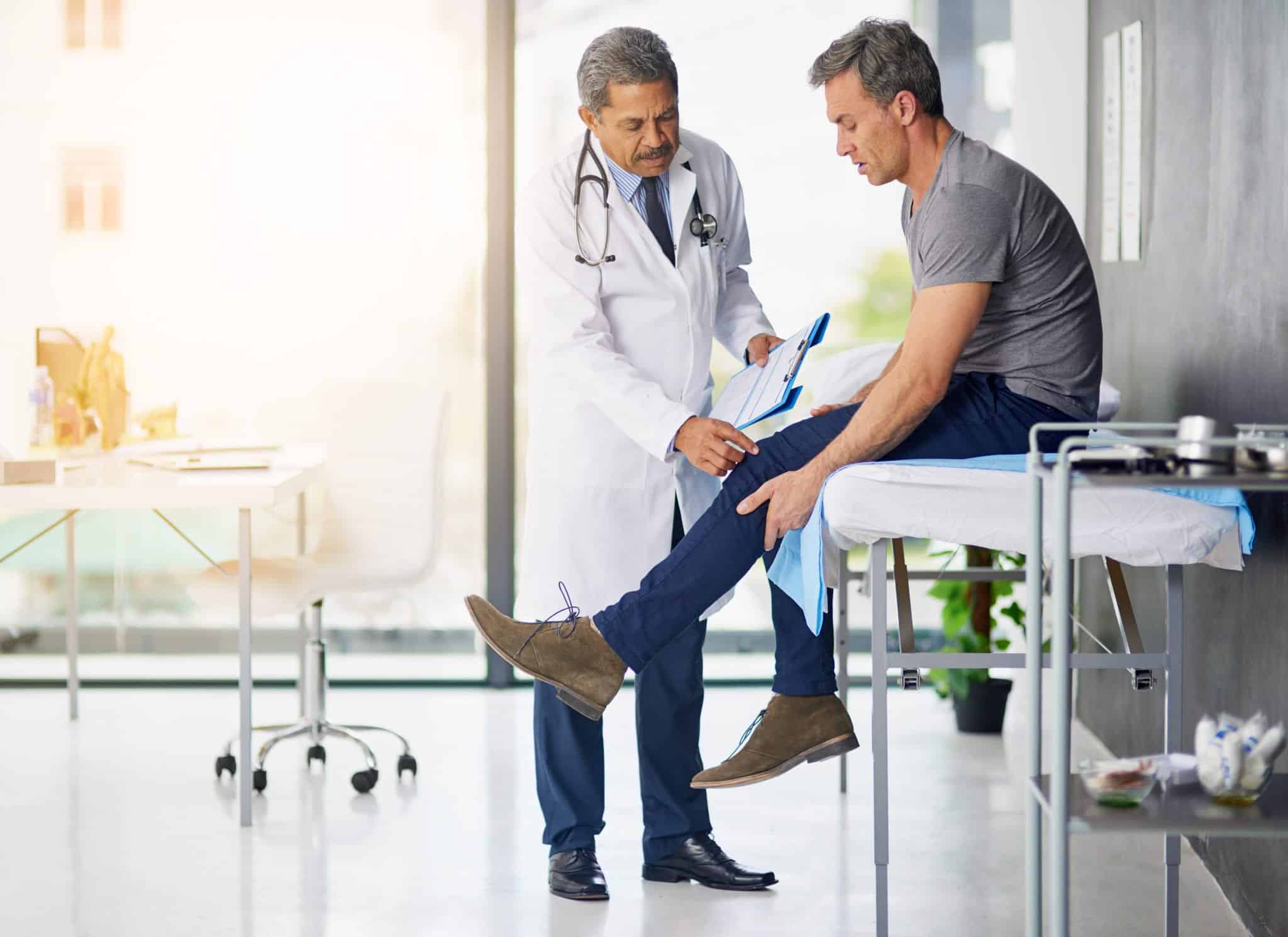 A male patient sitting on an exam table while a older male doctor examines his knee.