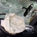 The inside of a wrecked vehicle with a shattered windshield, crocked rearview mirror and deployed airbag.