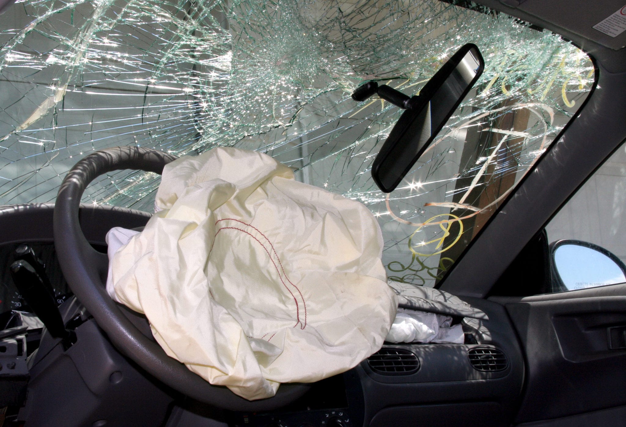 The inside of a wrecked vehicle with a shattered windshield, crocked rearview mirror and deployed airbag.