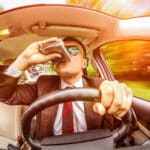 A view from behind a steering wheel of a car showing a man drinking out of a flask while driving.