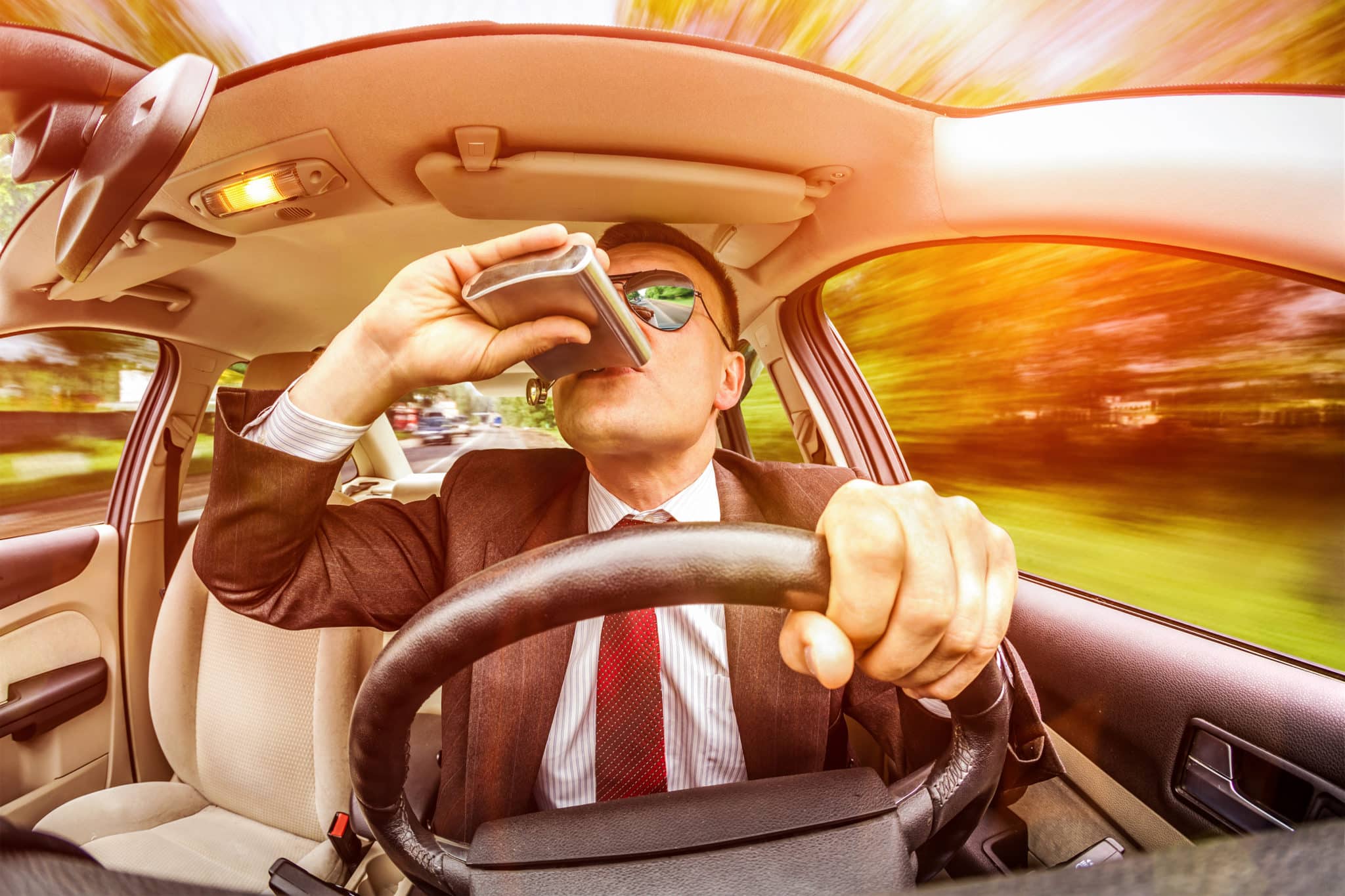 A view from behind a steering wheel of a car showing a man drinking out of a flask while driving.