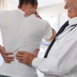 An older male doctor examining a male patient holding one hand on his neck and the other on his back.