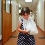 A Little girl with a white sling on her left arm, standing in the hallway of a hospital.