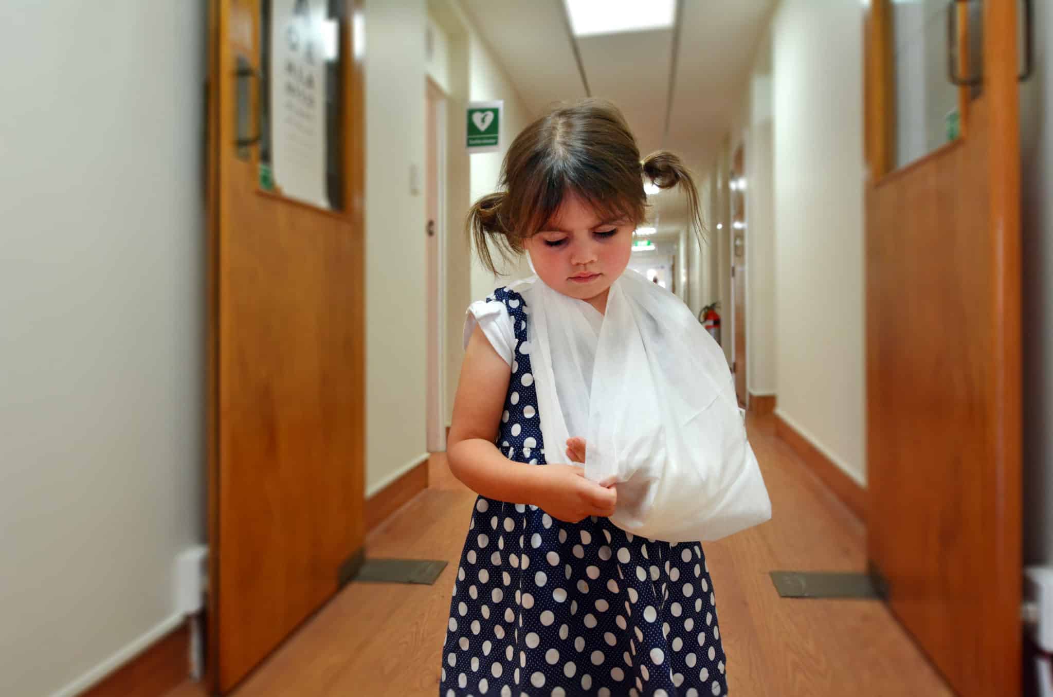 A Little girl with a white sling on her left arm, standing in the hallway of a hospital.