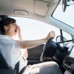 A tired woman driving a car with her right hand and covering her mouth with her left hand while she yawns.