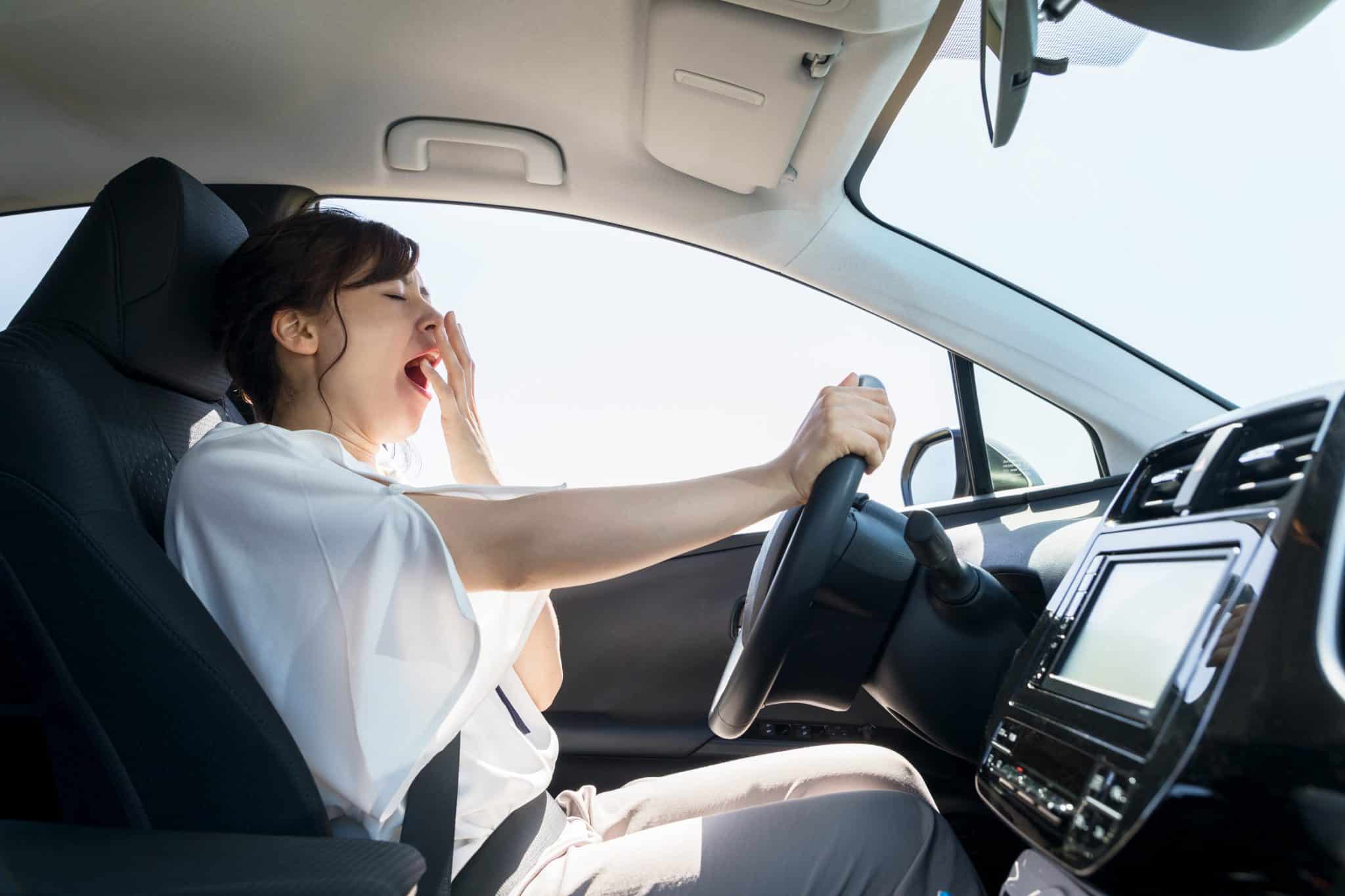 A tired woman driving a car with her right hand and covering her mouth with her left hand while she yawns.